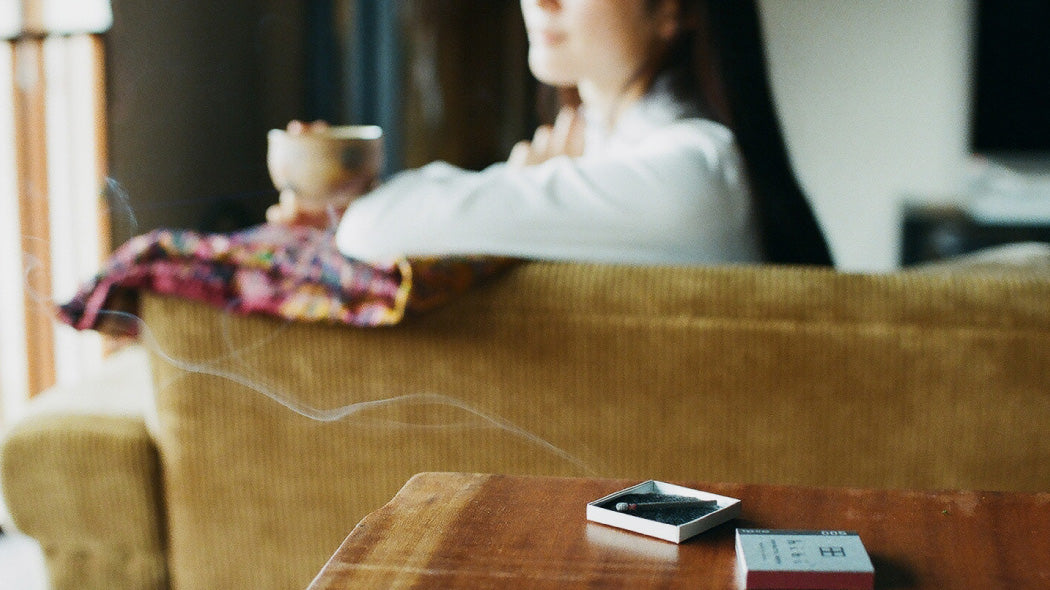 a hibi incense match is burning on its pad, a girl is drinking tea in the background