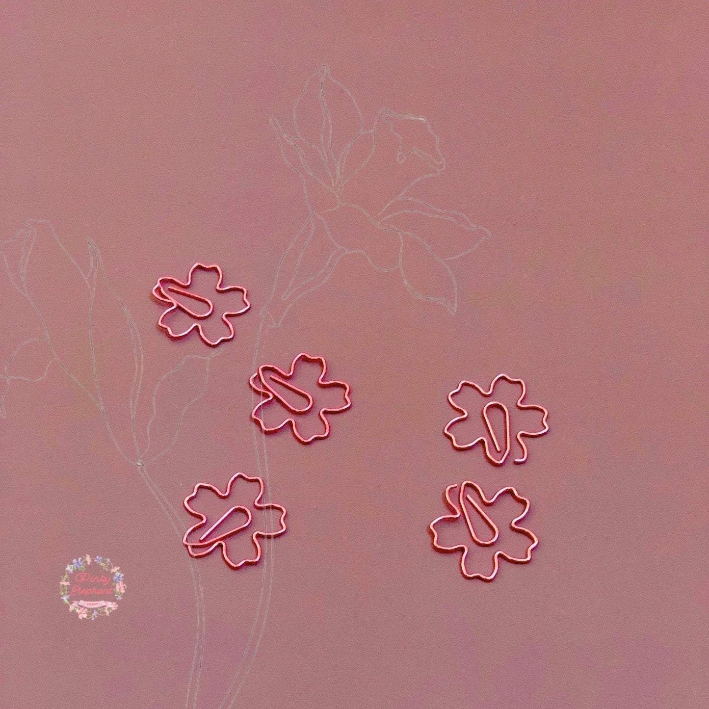 Five pink sakura-shaped paper clips on a light marron background, with a sketch of two roses in the background.