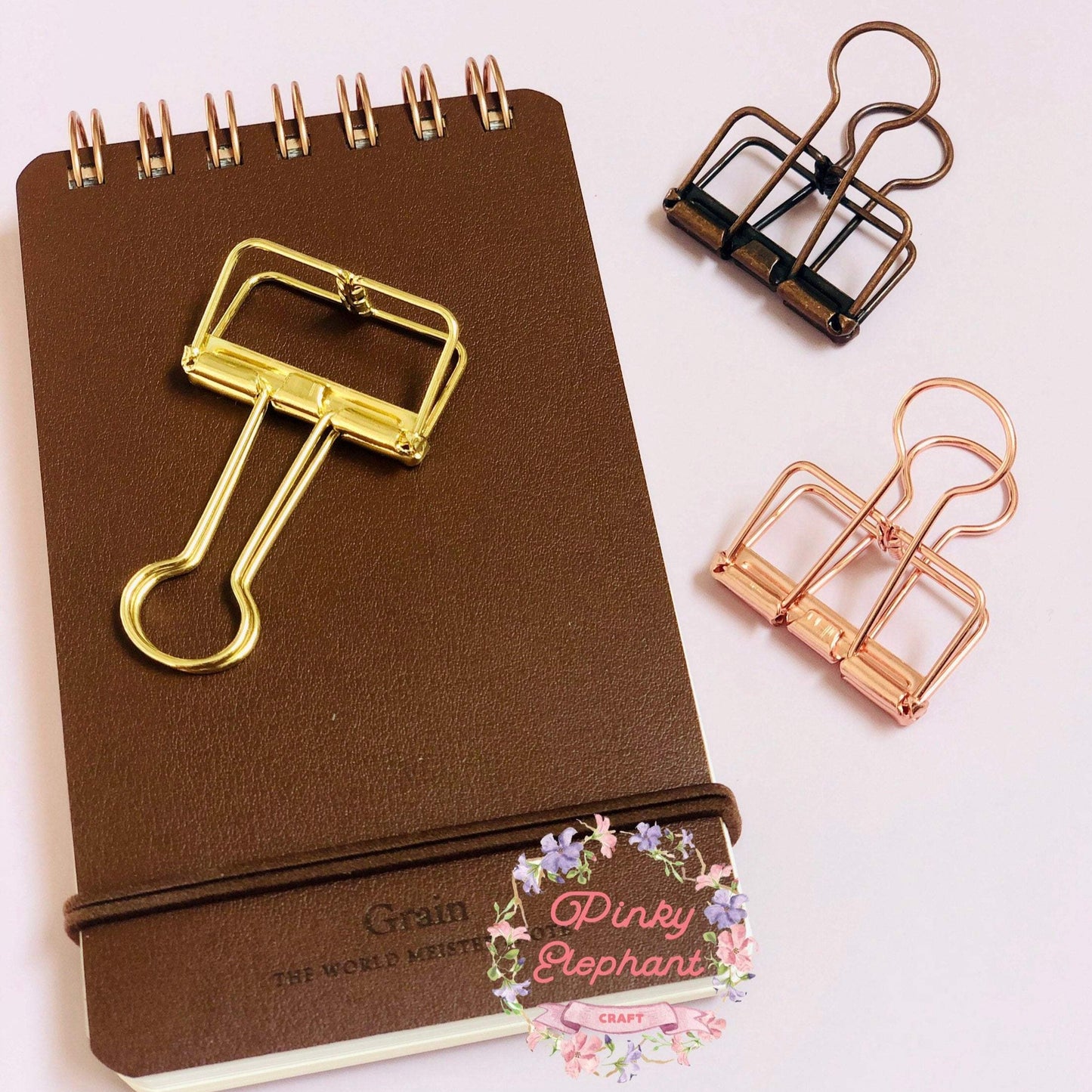 A golden skeleton paper clips placing on a flip leather cover memo pad, and there is a matte brown clip and a rose gold clip on their right. 