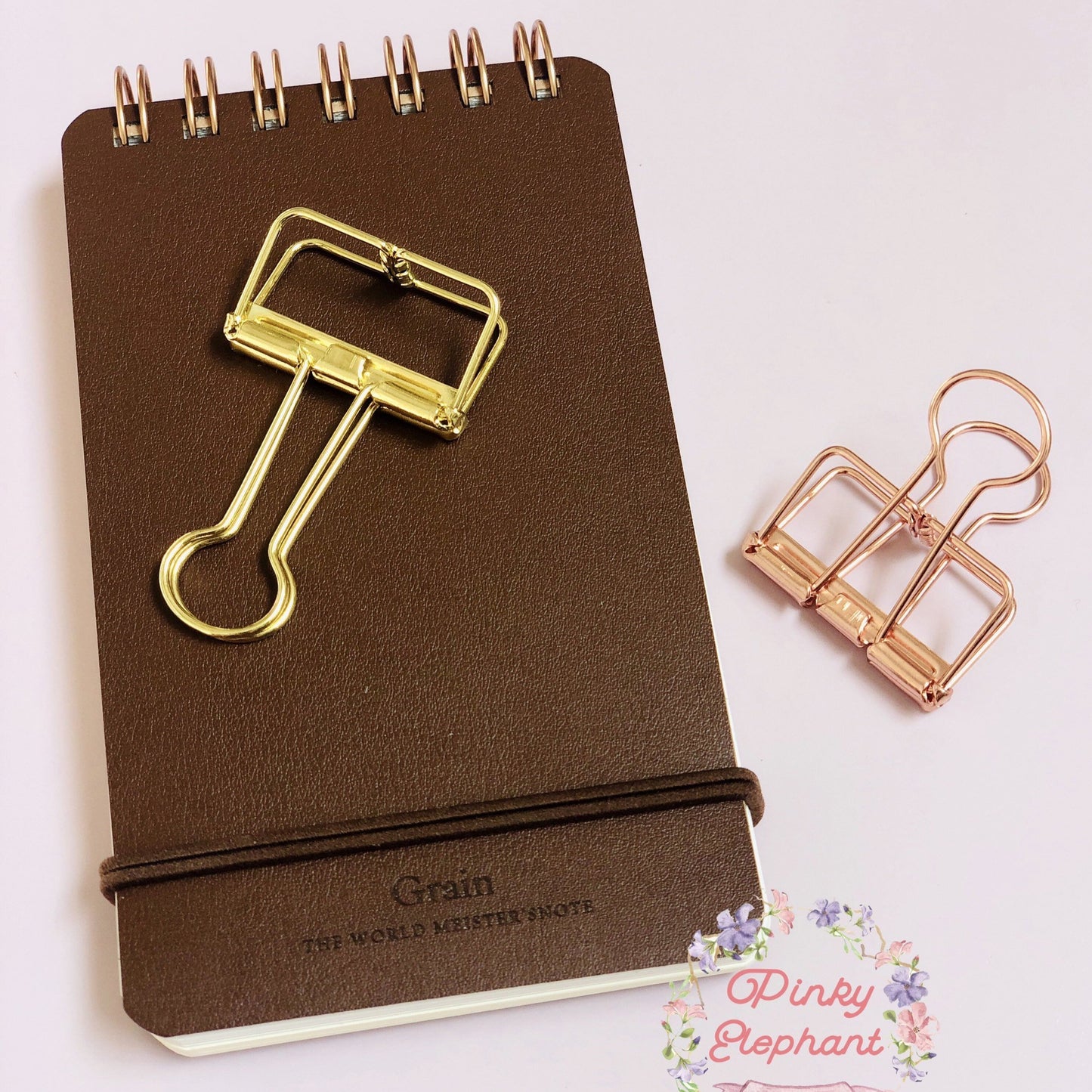 A golden skeleton paper clips placing on a flip leather cover memopad, and there is one rose gold clip on their right. 