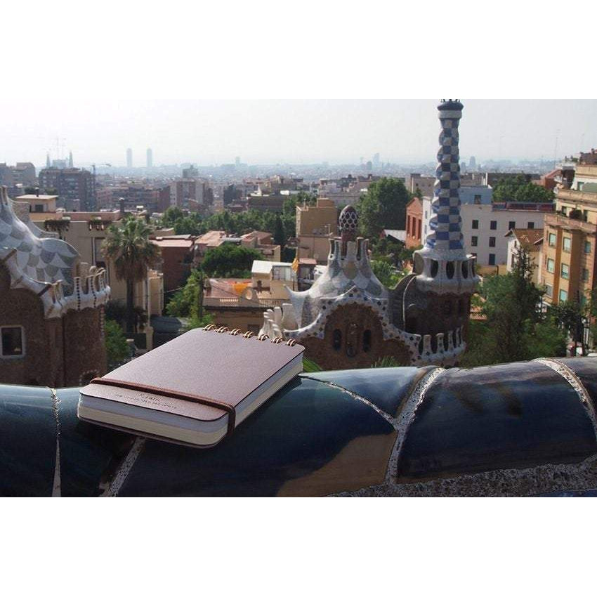 a Midori grain leather memopad in dark brown, placing on a balcony in Barcelona. Gaudí-styled buildings are in the background 