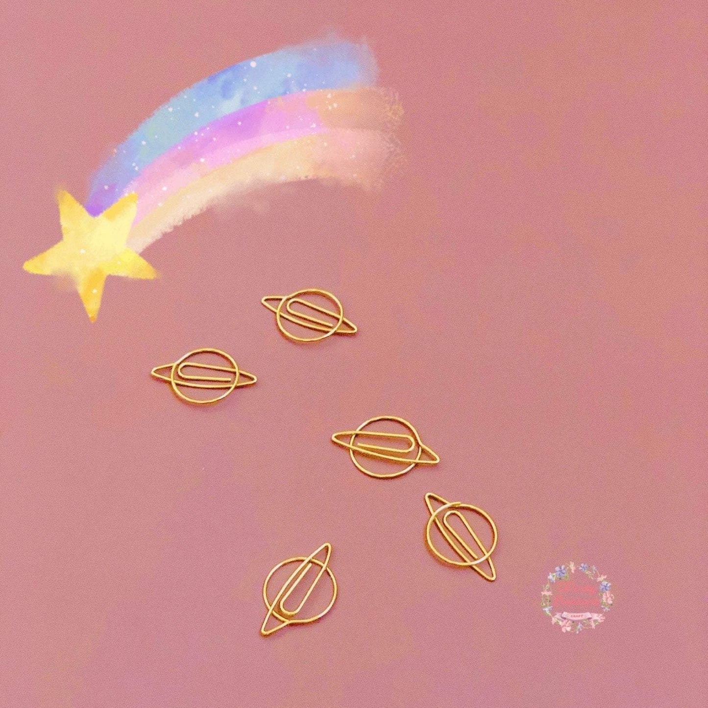 Five gold planet-shaped paper clips are in a pink background, A colorful meteor is drawn on the upper left