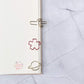 three clips clamping on a dotted notebook page, from the top to bottom, cactus-shaped, pink sakura-shaped and a gold planet-shaped, in a white marble background.