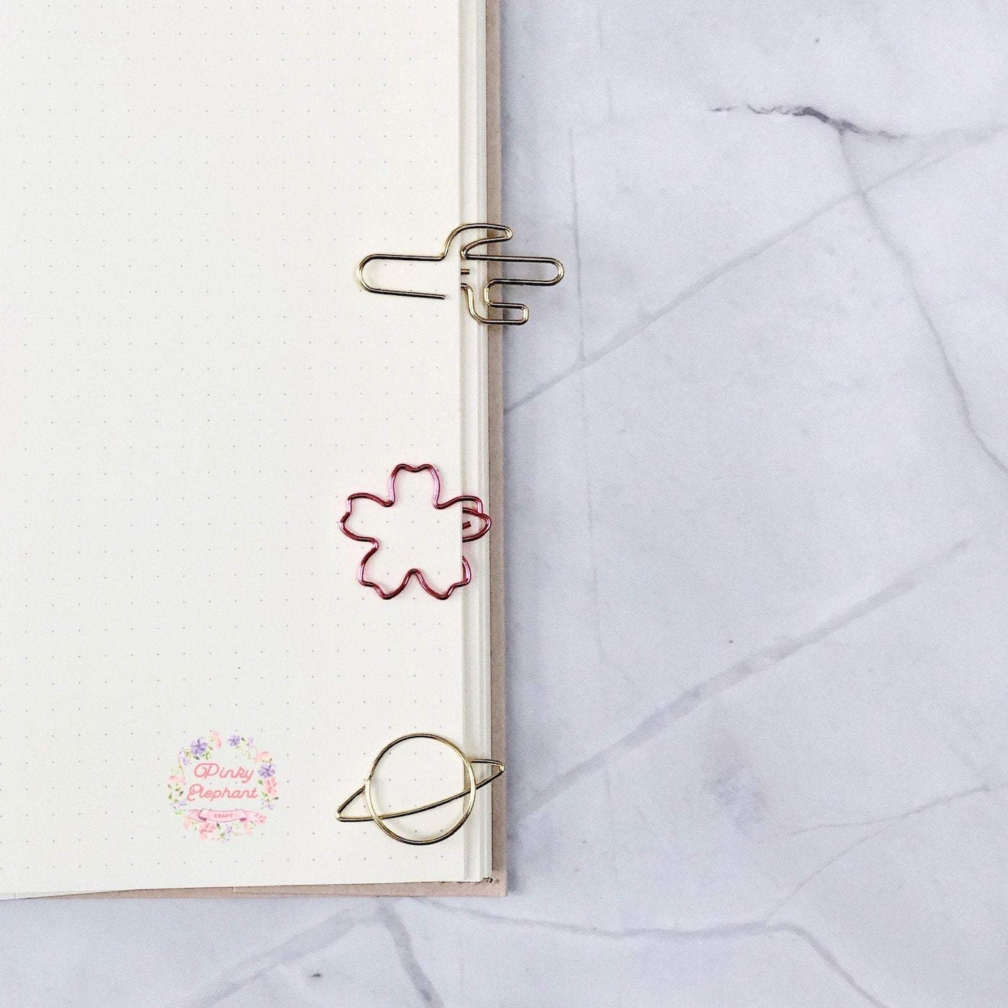 three clips clamping on a dotted notebook page, from the top to bottom, cactus-shaped, pink sakura-shaped and a gold planet-shaped, in a white marble background.