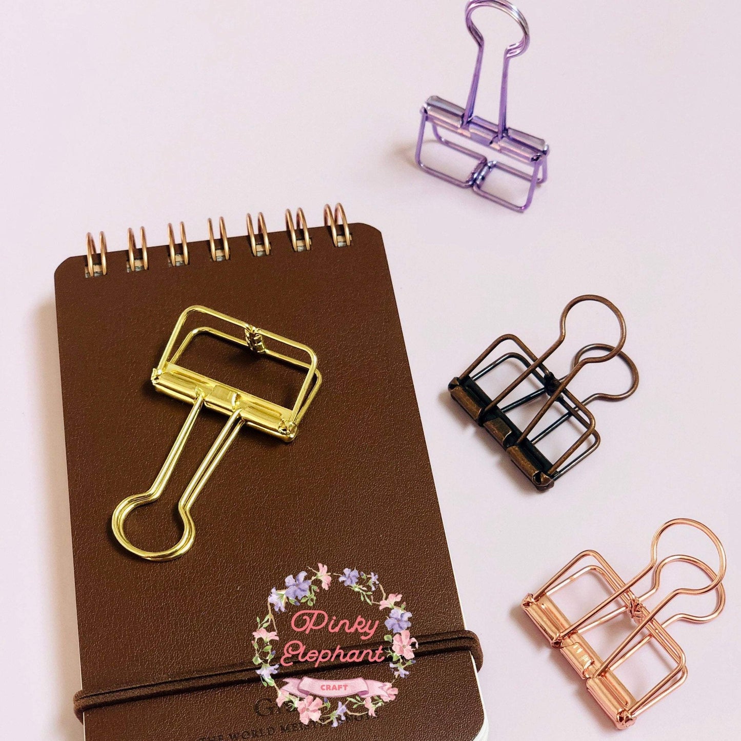 A golden skeleton paper clips placing on a flip leather cover memopad, there are a metallic  purple clip, one matte brown clip and one rose gold clip on their right.