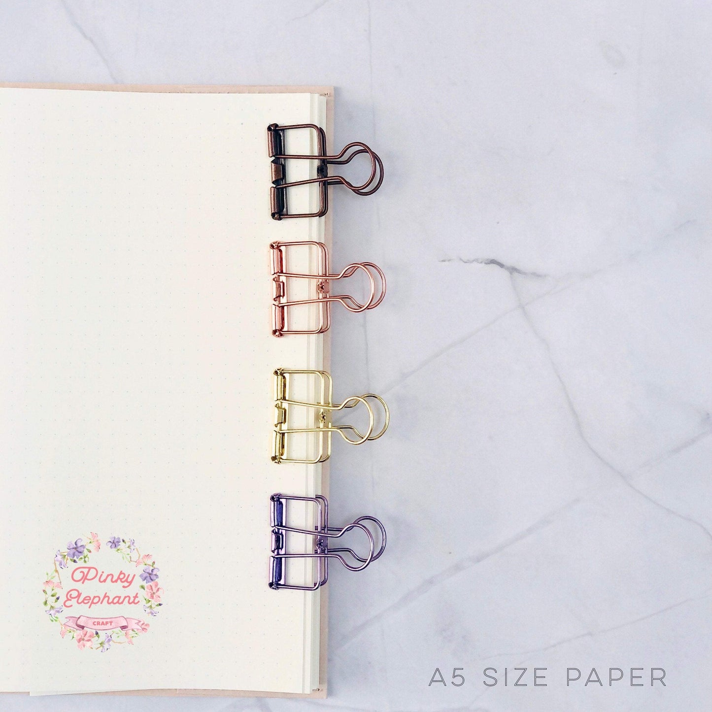 Four skeleton clips are clamped on the open A5 dotted notebook. Their colors from top to bottom are matte brown, rose gold, gold and metallic purple. 